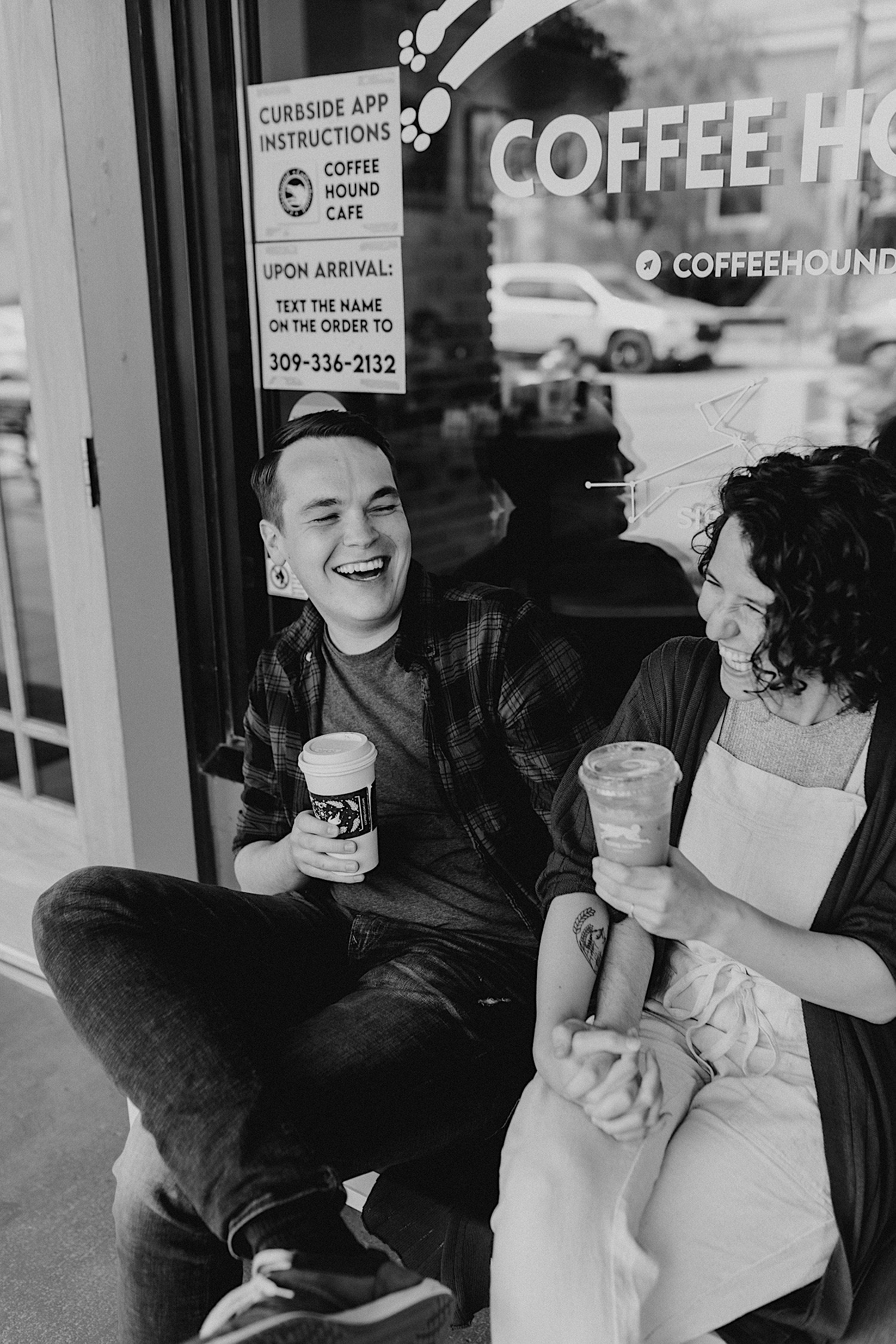 A black and white photo of a couple laughing during their engagement session in Normal Illinois outside a coffee shop