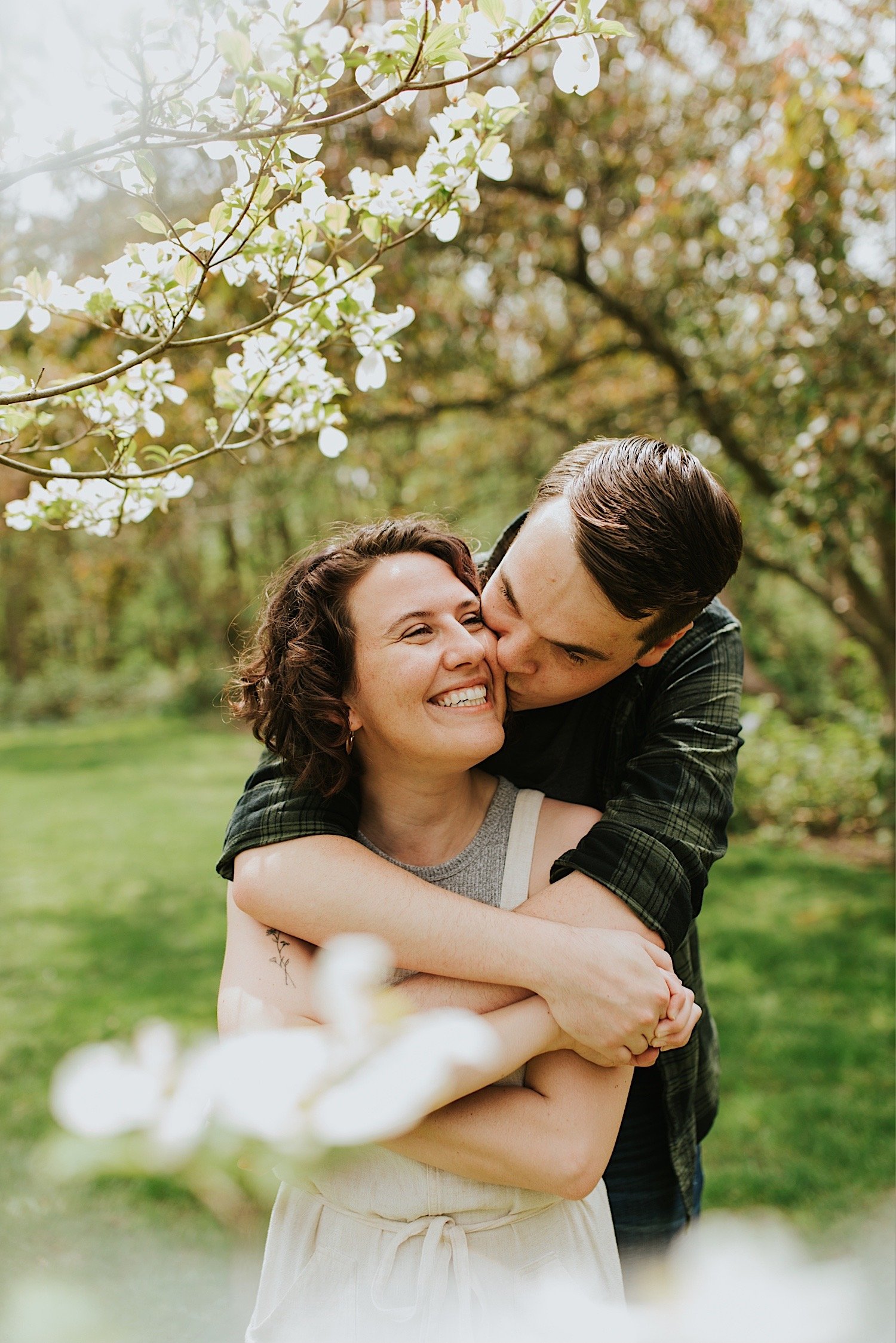 future groom holds his future bride under a tree with white flowers in a park in Normal Illinois as he kisses her cheek