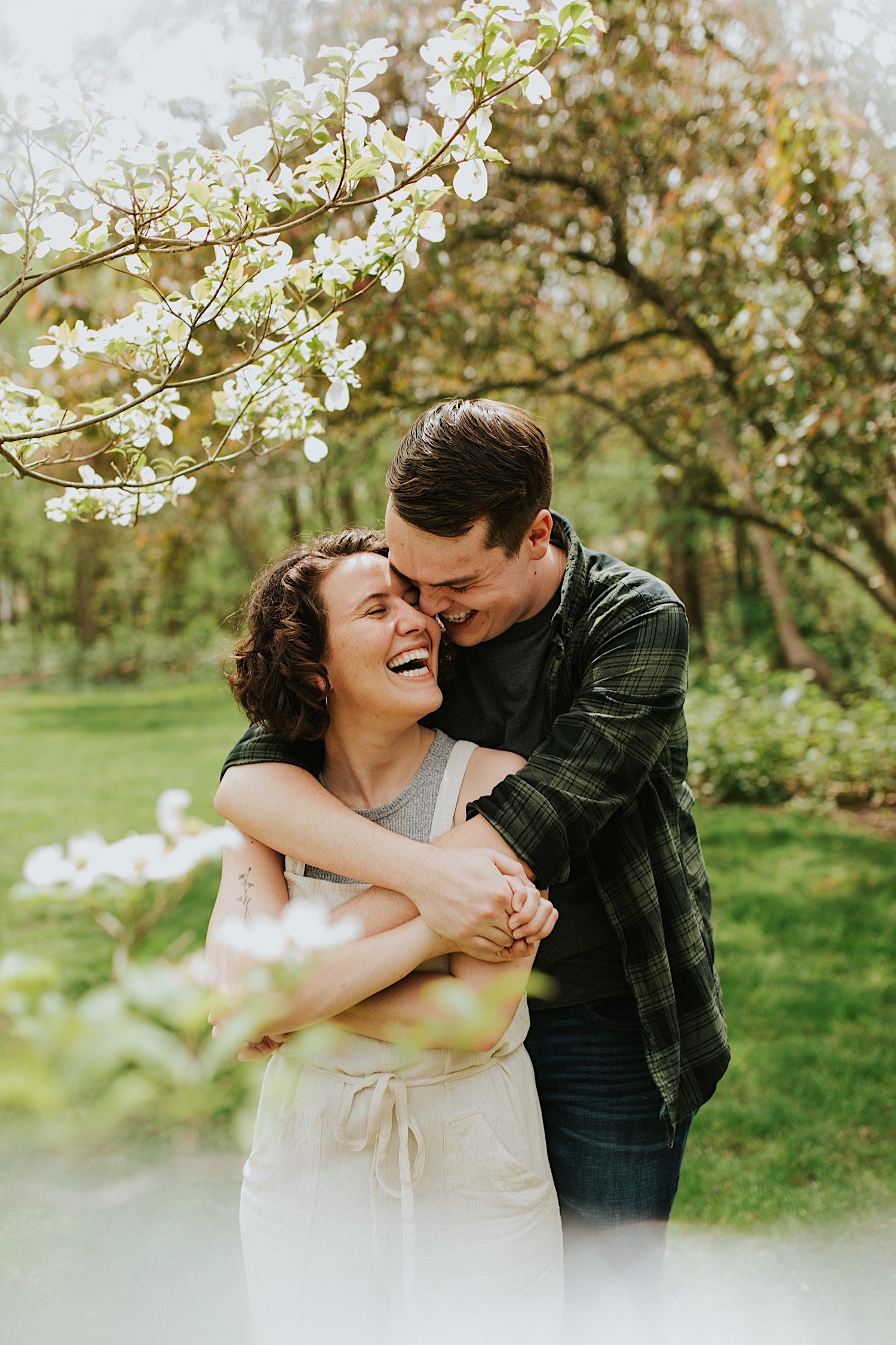 future groom holds his future bride under a tree with white flowers in a park in Normal Illinoisas they both laugh