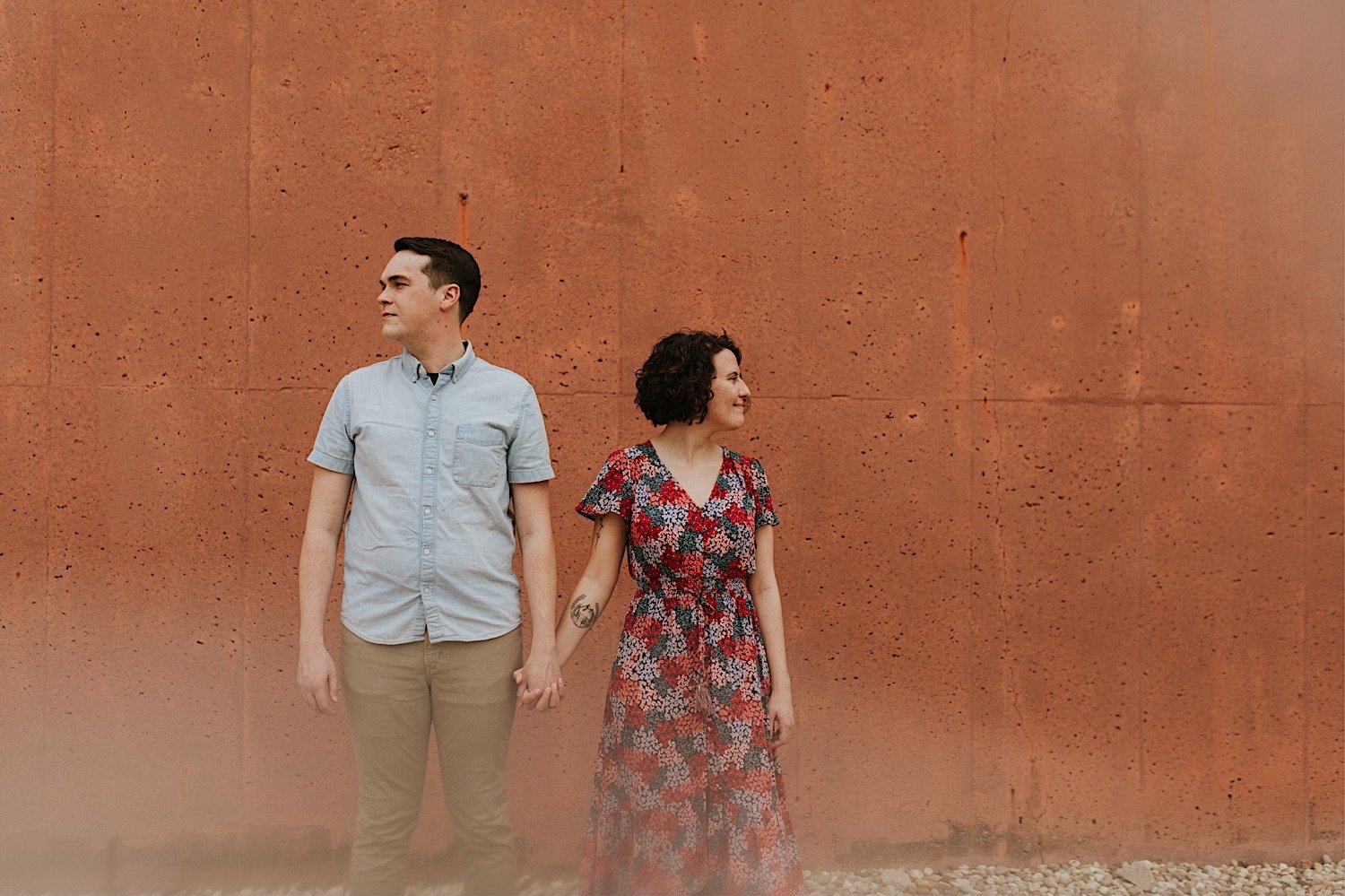A couples holds hands showing off their outfits during their engagement session in Normal Illinois.  She wears a floral dress and he is wearing a blue short sleeved button up and khakis for their engagement session