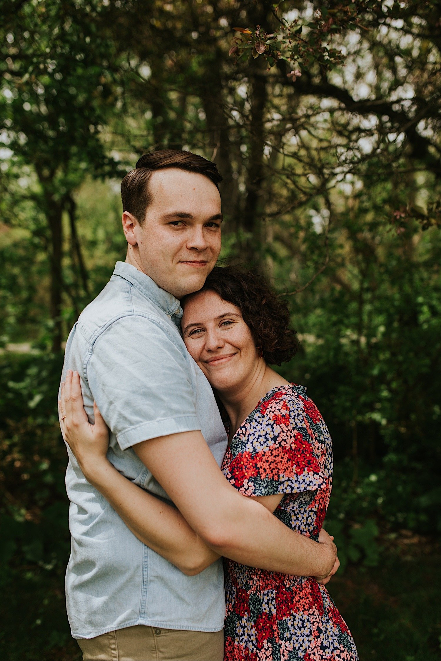 A couple hugs while smiling at the camera while standing in front of a forest in Normal Illinois