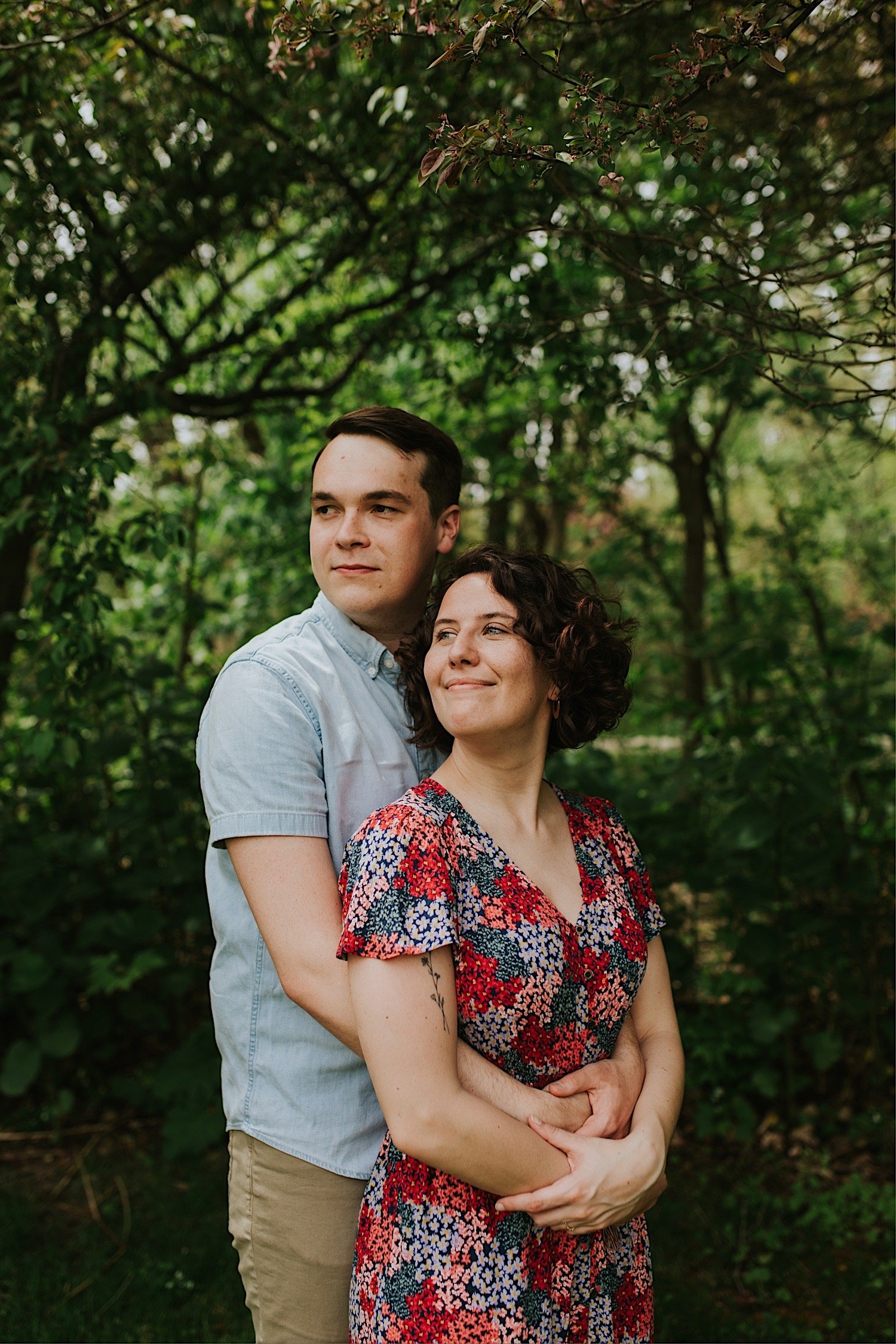 A couple hugs while smiling at the camera while standing in front of some trees at a park in Normal illinois