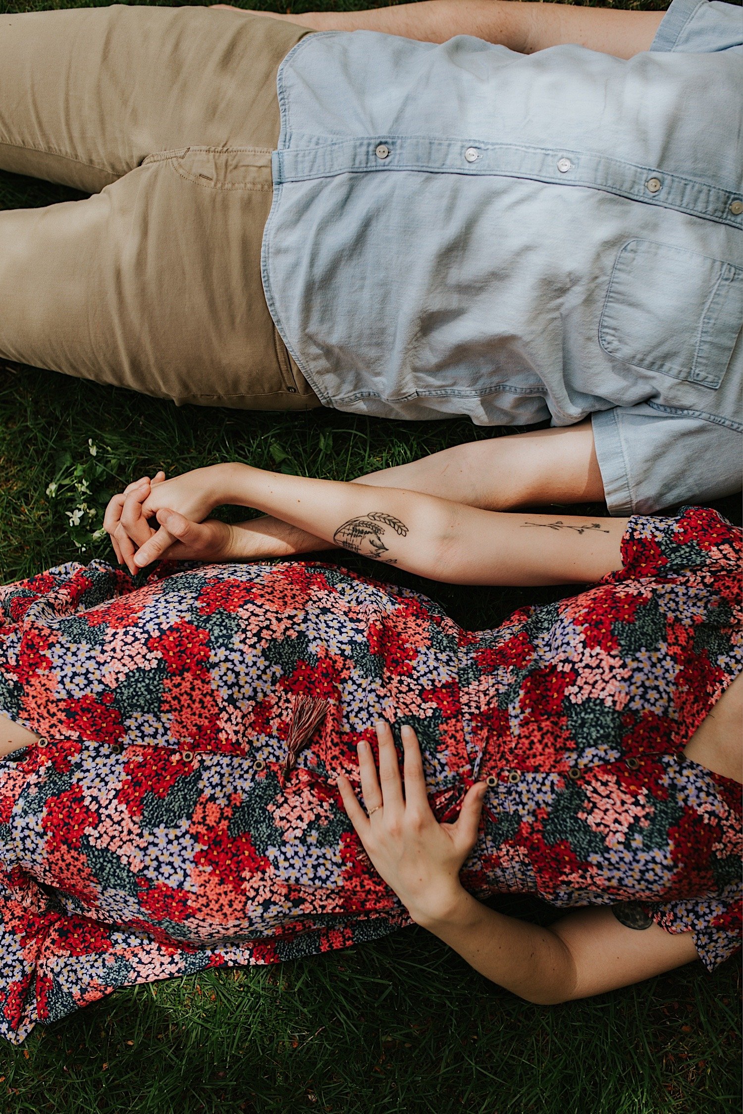 An engaged couple lays in the grass and holds hands during their engagement session.  The image is of their torsos and hands holding one another, showing off their spring time outfits