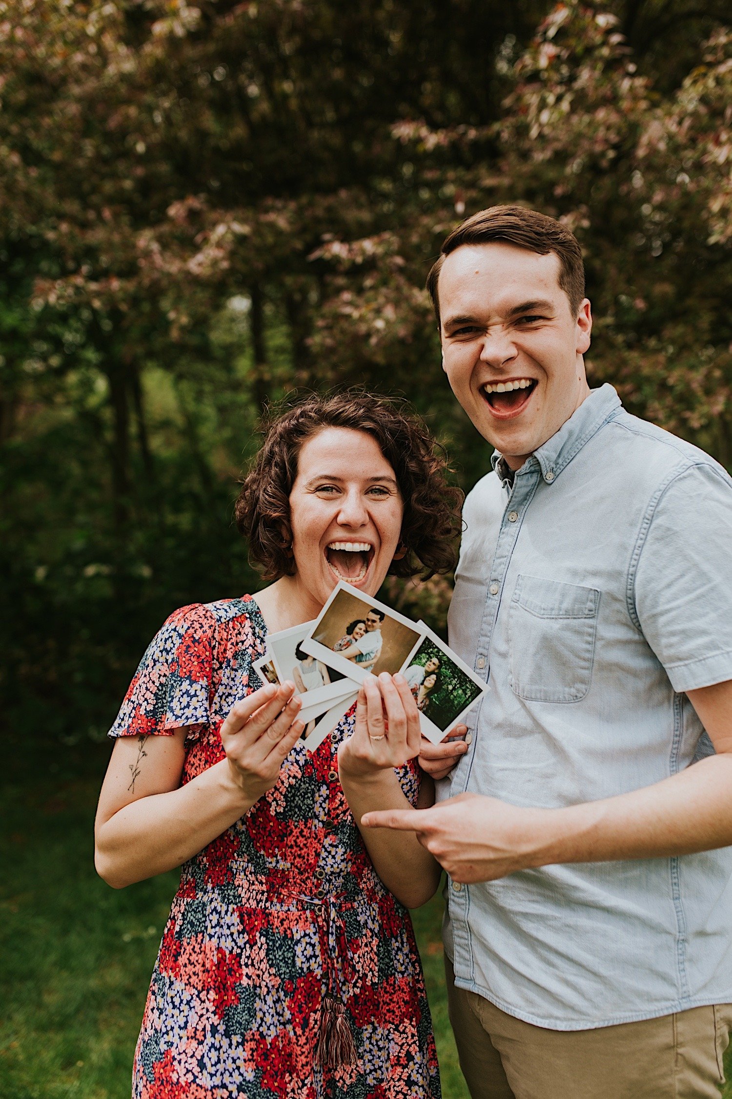 An engaged couple smiles at the camera while holding polaroid photos of themselves from their engagement session