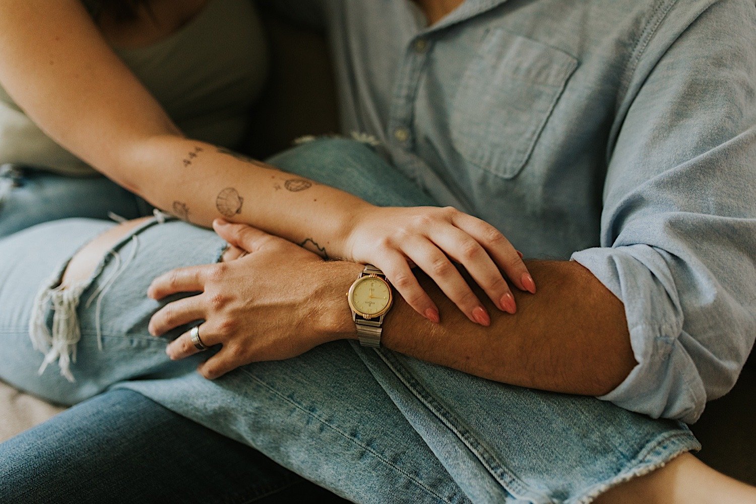 A close up of a woman's hand holding her husbands arm.  They are sitting on the couch with their legs overlapping.  The husband has a gold watch on with a silver strap and a silver wedding band.  The wife has a shell tattoo on her arm.