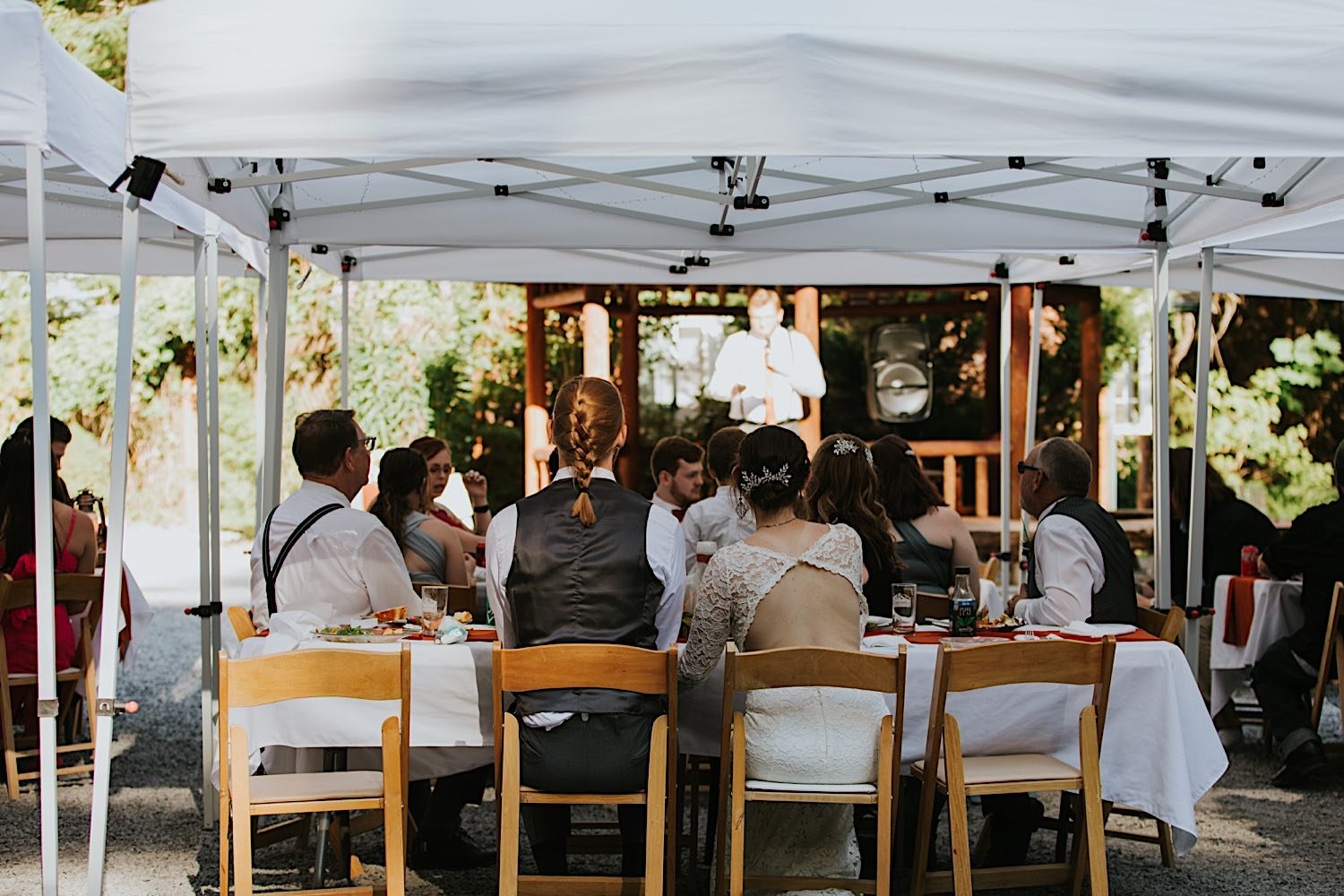 A newly married couple sits in wooden folding chairs underneath a pop up tent at their intimate wedding during a speech.