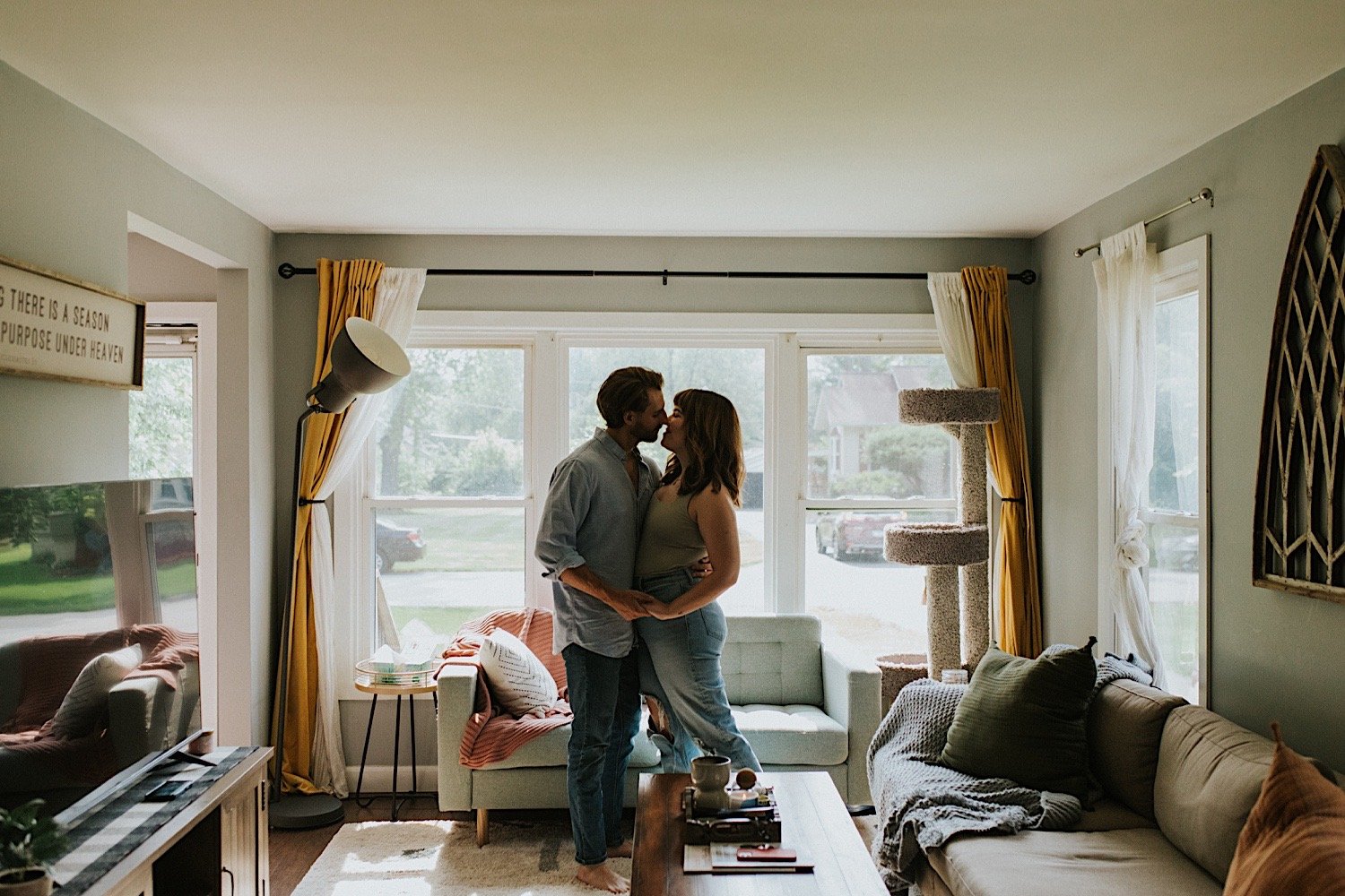 A married couple holding hands and kissing while standing in front of the window in their living room.