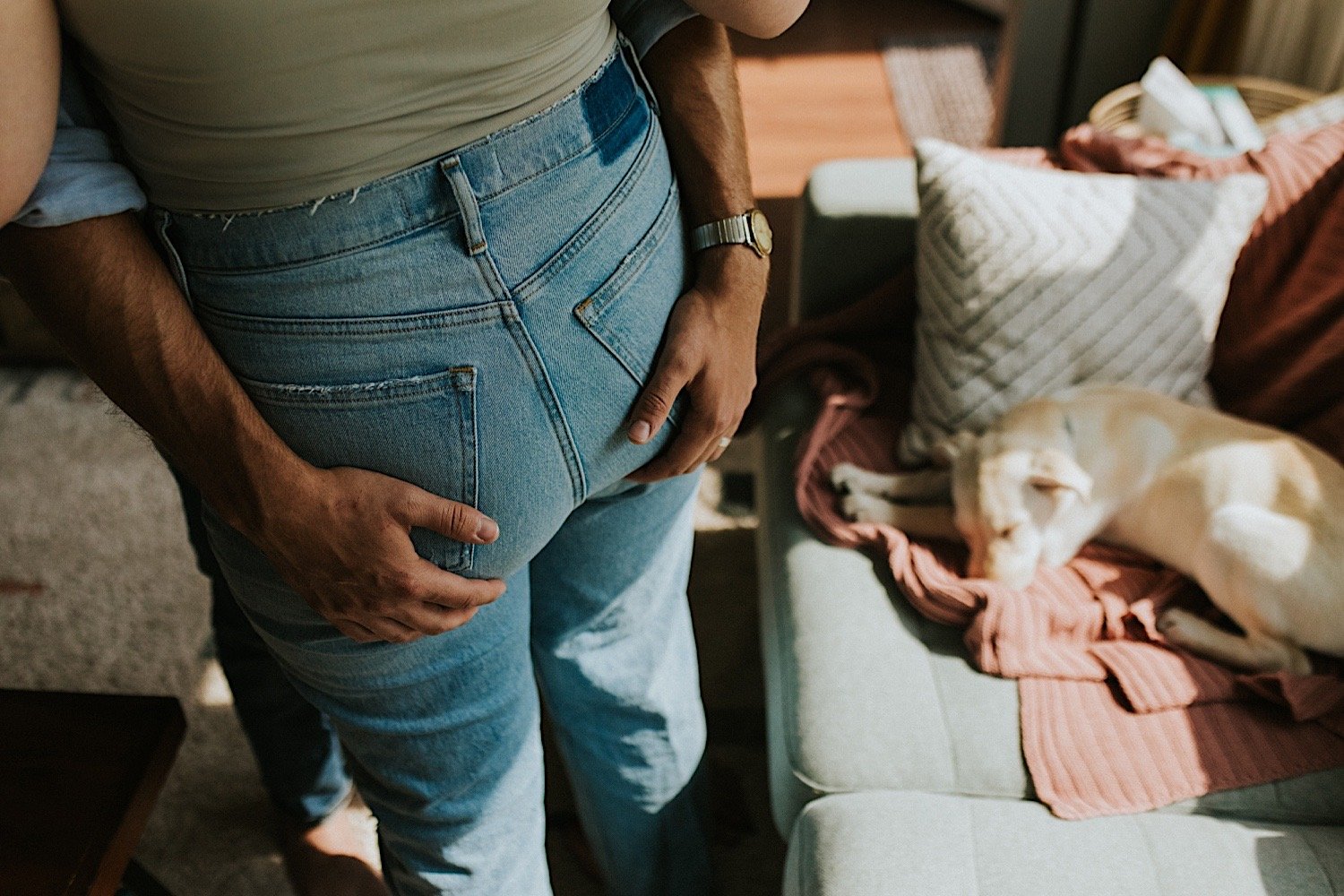 A close up image of a husband and wife hugging tight, the husband grabs his wife over her back denim pockets.  There is a pup sleeping on the couch in the background.
