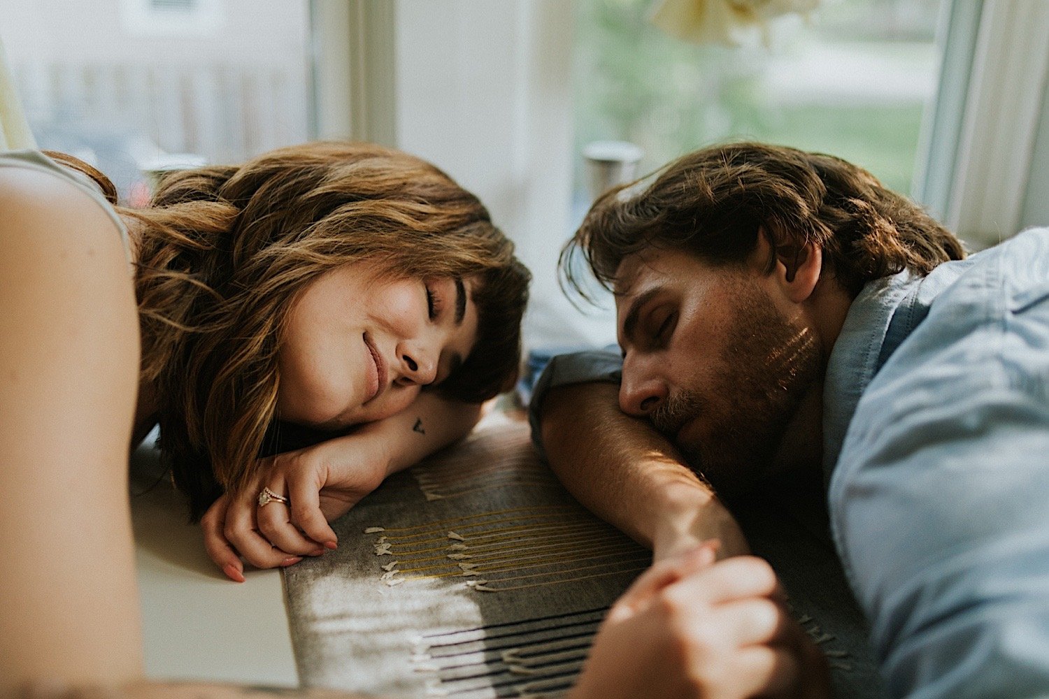 A close up image of a husband and wife lay their heads on their kitchen table with their eyes closed.  They are holding hands.
