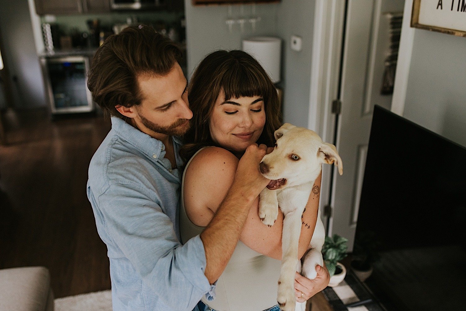 A wife holds her puppy while her husband stands behind her hugging them both.