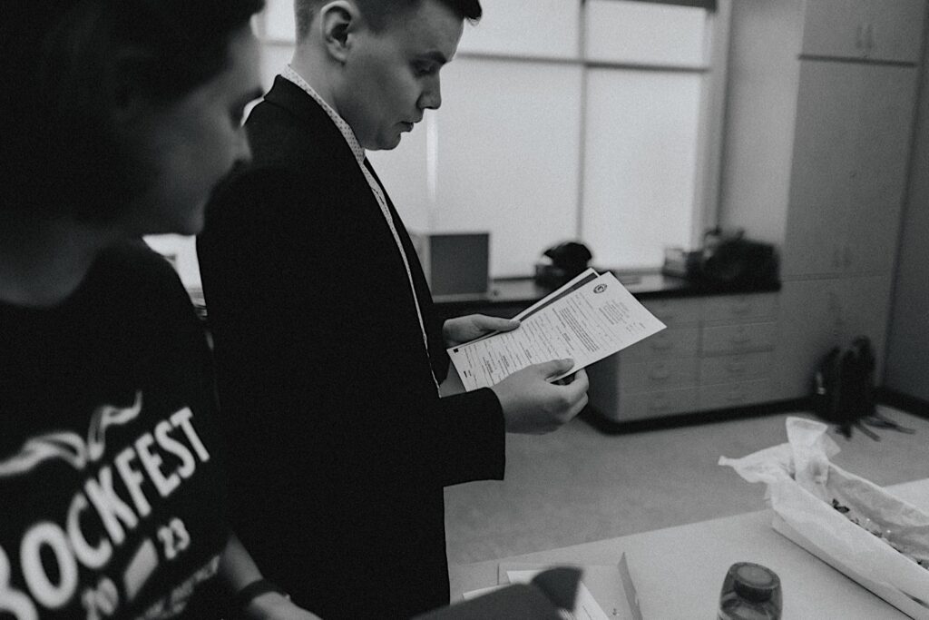 Black and white photo of a groom reading over the paperwork for his wedding with a friend by his side