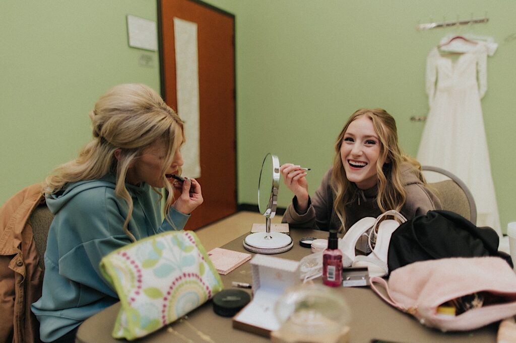 Two women sit at a table with a small circle mirror on it at they put on their make up before a wedding, the girl on the right is smiling at the camera