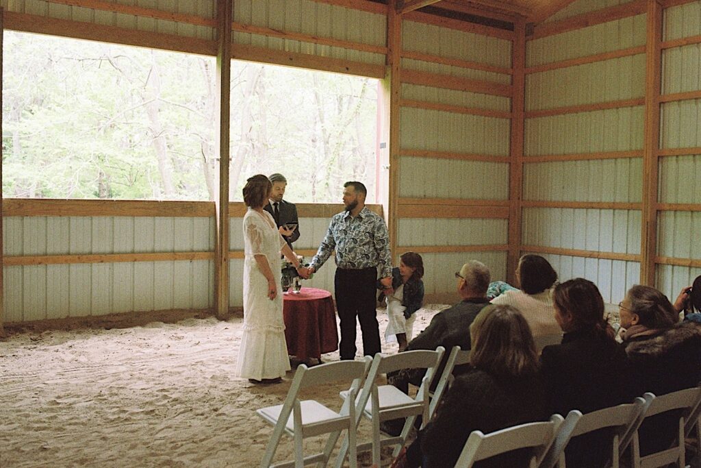 Film photo of a bride and groom during their wedding ceremony in their barn, with guests seated watching and the groom's daughter holding his hand
