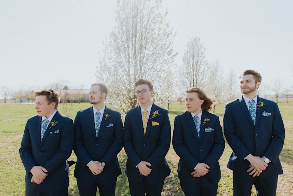 A groom stands with his four groomsmen and looks at the camera while they all look to the left while standing in a field