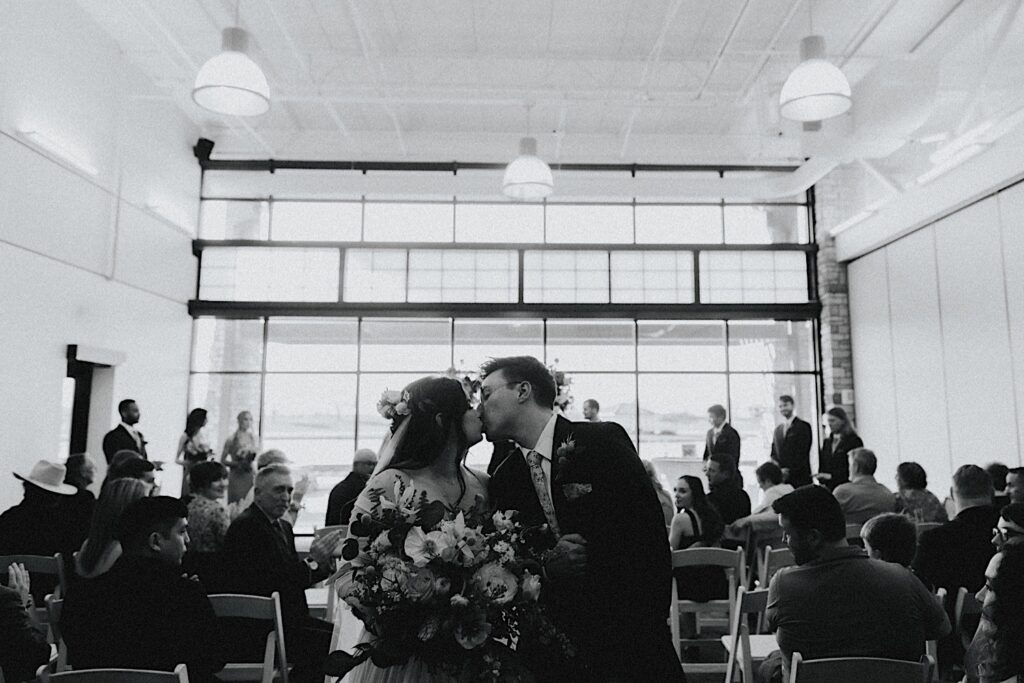 Black and white photo of a bride and groom kissing while walking down the aisle after their indoor wedding ceremony at Erin's Pavilion