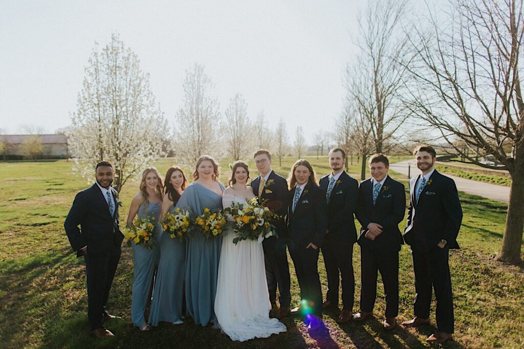 A bride and groom stand with their wedding parties on either side of them and all smile at the camera in a field outside Erin's Pavilion