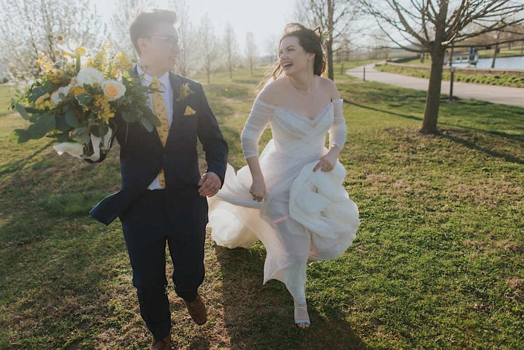 A bride and groom smile as they walk side by side with one another in a field outside Erin's Pavilion during sunset, the groom is carrying the bouquet of yellow flowers