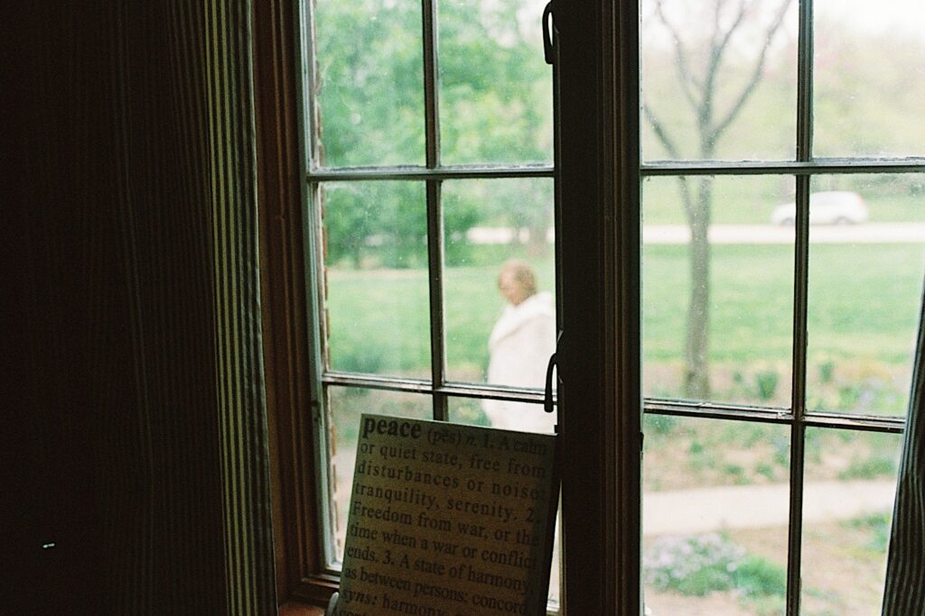 Film photo of a sign that reads "Peace" followed by the definition, the sign sits in front of a window and outside the window is the bride standing in the front yard