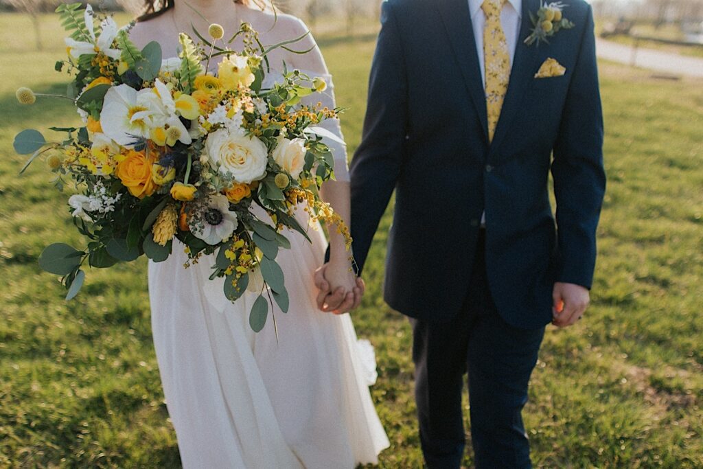 Shoulder down photo of a bride and groom holding hands while standing in a field, in the bride's other hand is her yellow bouquet