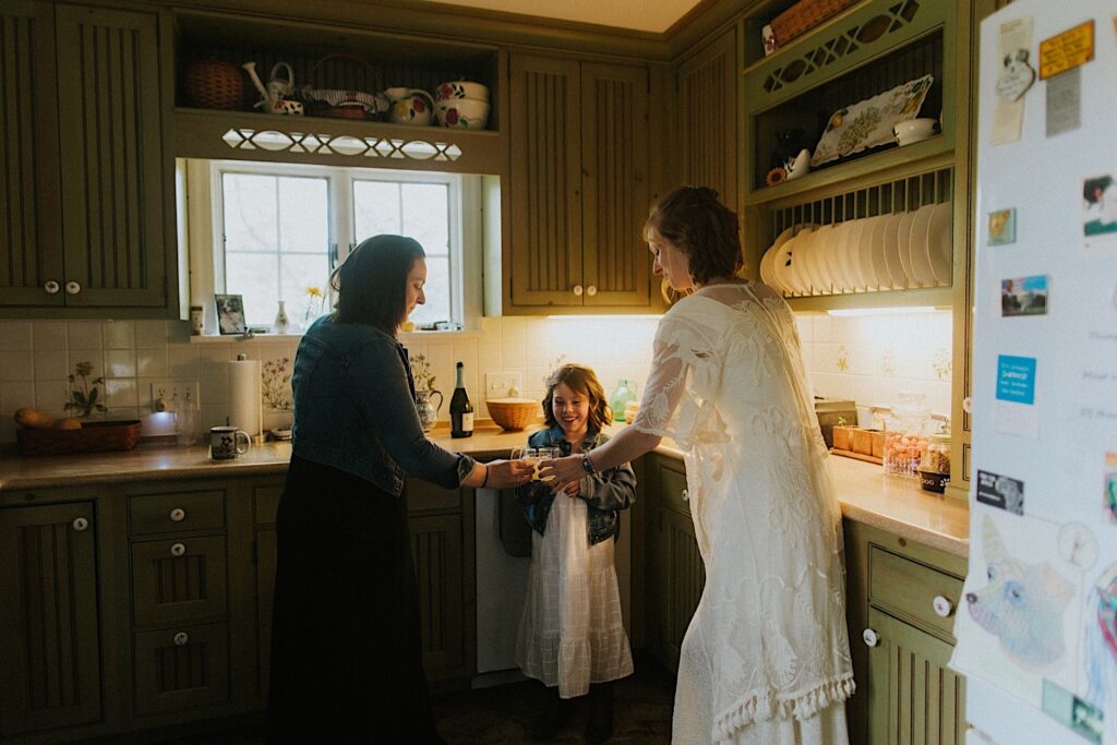 A bride stands in a kitchen with a woman and a young girl, each of them has a glass and they are toasting them