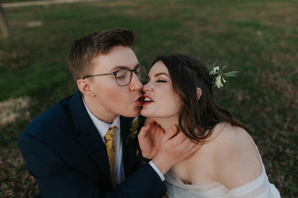 A bride and groom sit in the grass and are about to kiss while making funny faces