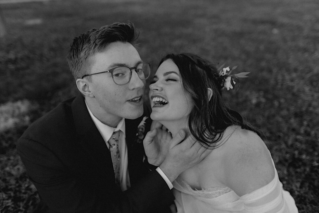 Black and white photo of a bride and groom sitting in grass, they were leaning in for a goofy kiss and the groom is saying something while the bride laughs