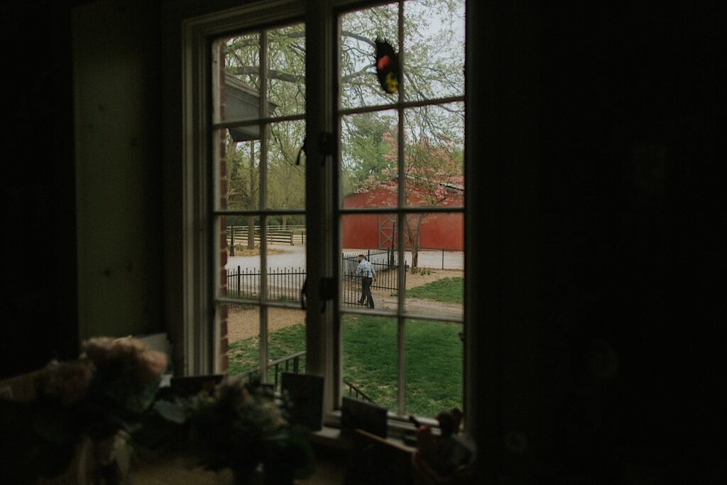 Photo taken inside a house of a window, outside the window a man walks through a metal gate next to a barn