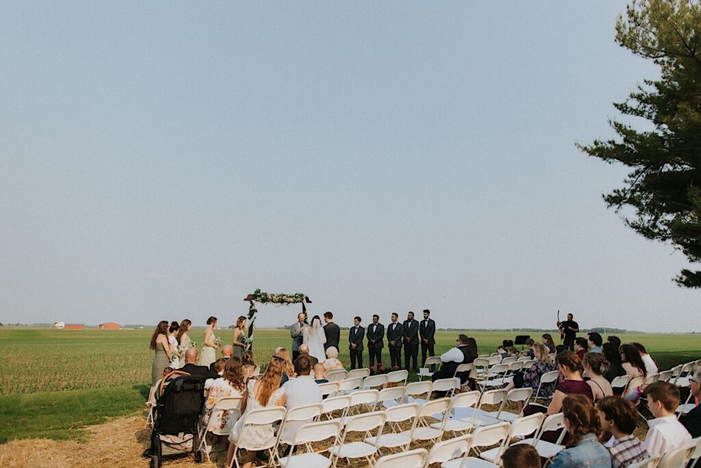 A wedding ceremony is taking place in a field, chairs are set up looking out at the ceremony