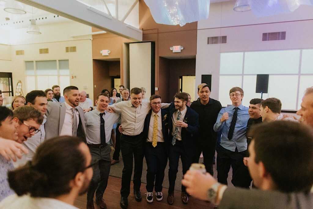The groom stands in a circle with a bunch of other men with their arms on each other's shoulders as they dance during the wedding reception at Erin's Pavilion