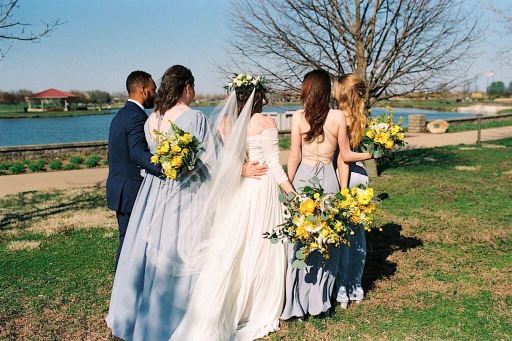Photo of a bride and her 4 members of her wedding party embracing and facing away from the camera looking at a nearby lake