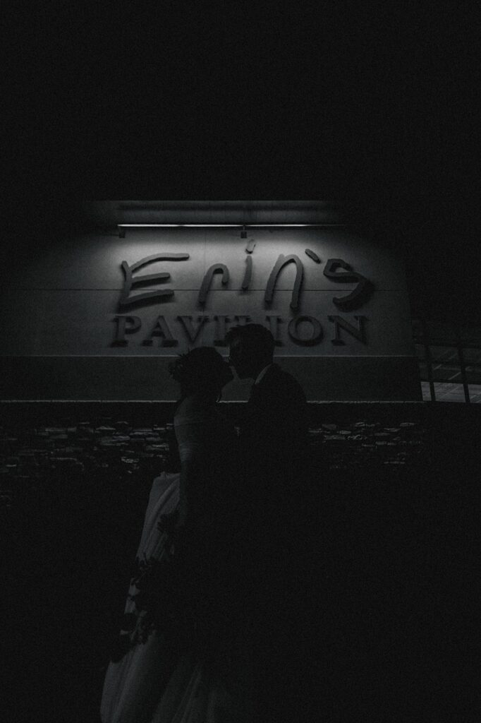 Black and white photo of a bride and groom about to kiss, behind them is a building with a sign reading Erin's Pavilion above them