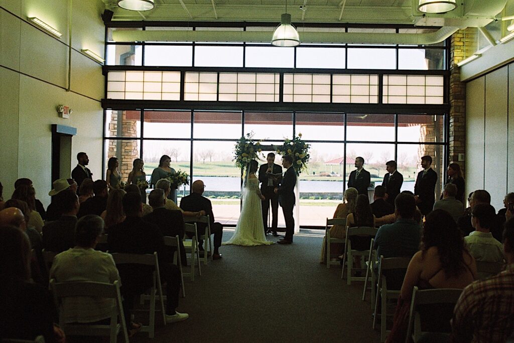 Film photo of a bride and groom during their indoor wedding ceremony as guests sit and watch and the officiant performs the service, taken by a documentary wedding photographer