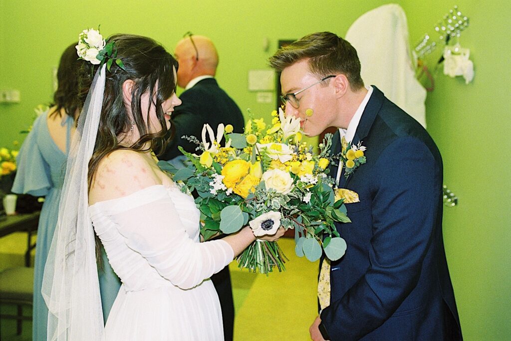 Film photo of a bride holding out her bouquet to the groom who is smelling it