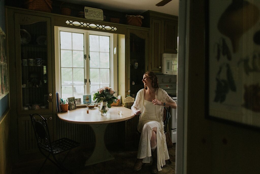 A bride sitting in a kitchen laughs as she looks out the window before her backyard wedding in Springfield Illinois