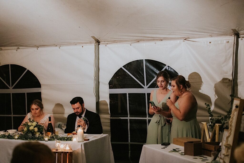 A bride and groom sit at a table and eat during their tent wedding reception while two bridesmaids to the right of them read from a phone while giving a speech 