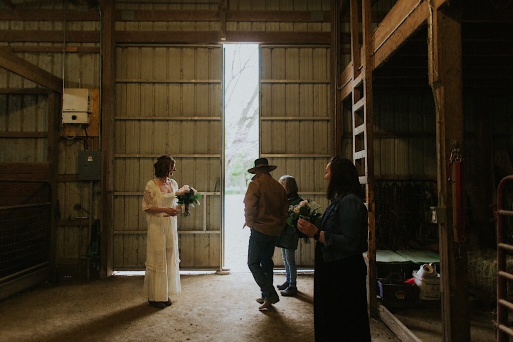 A bride stands in a barn next to three other guests of their backyard wedding in Springfield, one man in a cowboy hat is walking towards the open door of the barn