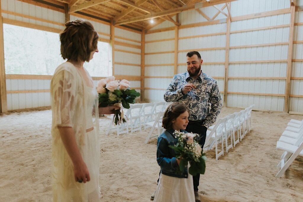 A bride and groom stand in a barn that is set up as their ceremony space for their backyard wedding in Springfield Illinois, between them is a young girl holding a bouquet