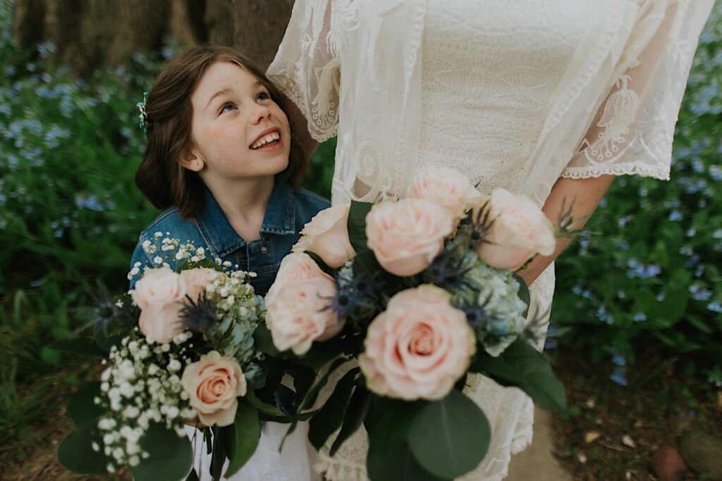 A young girl looks up and smiles at a bride who is standing next to her, both of them are holding bouquets