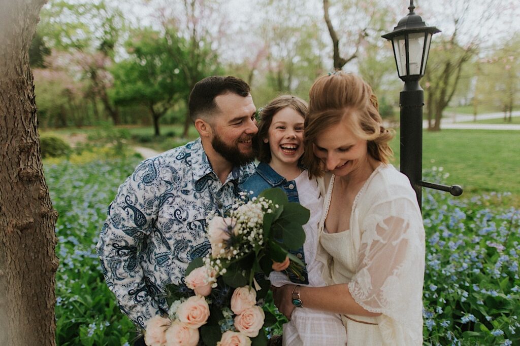 A bride and groom hold their daughter between them as they all laugh and smile during portraits in their backyard for their wedding day in Springfield Illinois