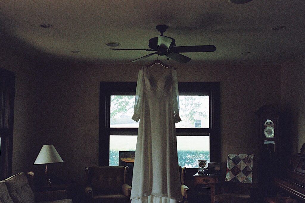 Film photo of a wedding dress hanging in a living room from a ceiling fan, taken by a documentary wedding photographer