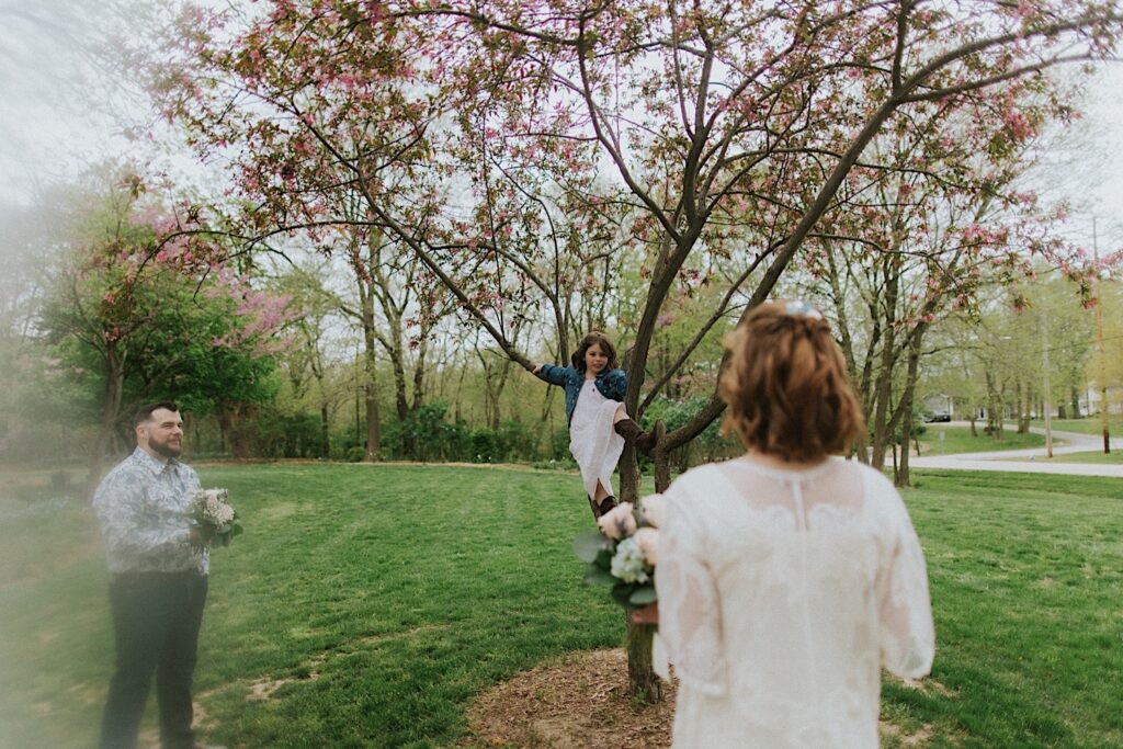 A bride and groom look on as their young daughter climbs a tree in front of them in their backyard