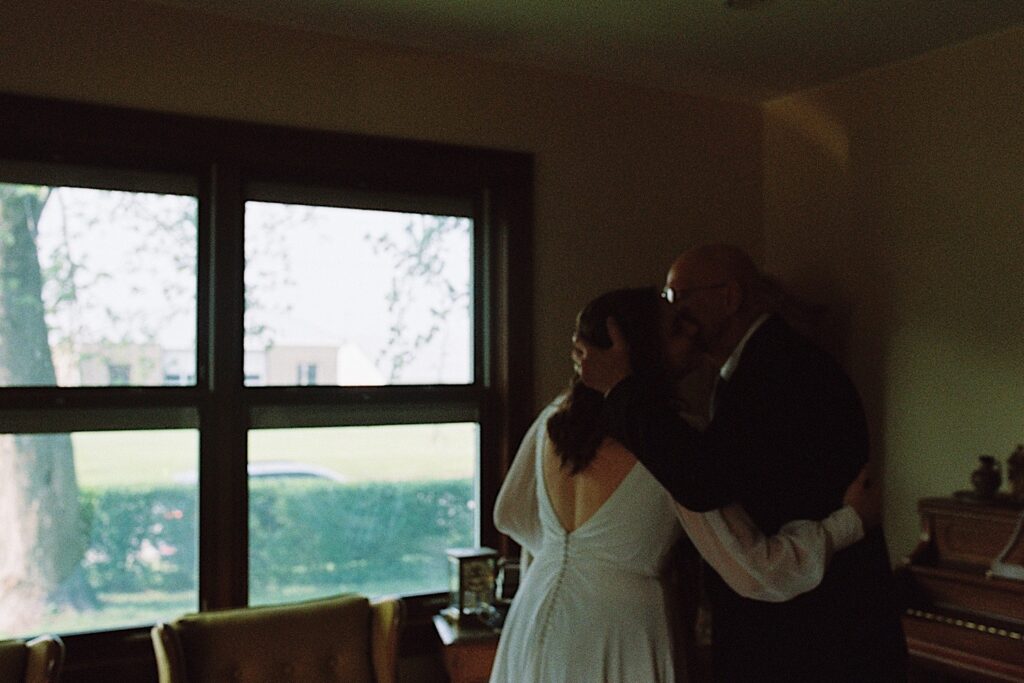 Film photo of a bride embracing her father in the living room as he kisses her on the cheek, taken by a documentary wedding photographer