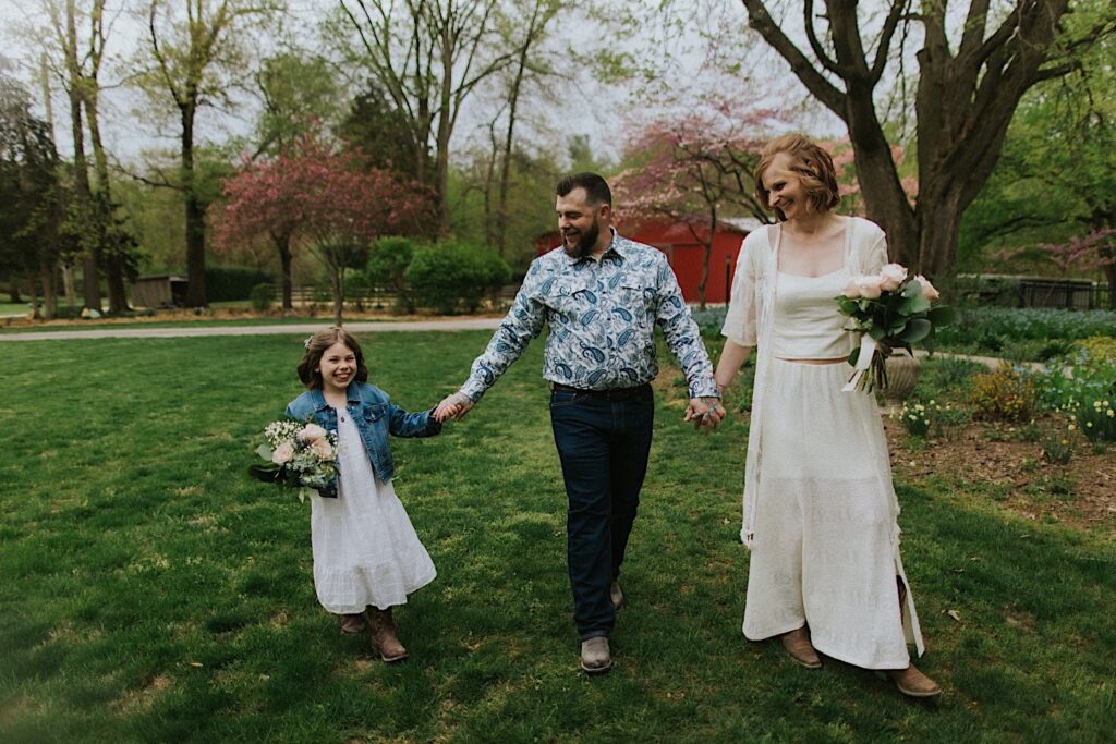 A bride and groom walk with their daughter as they all hold hands and smile, they're in their backyard taking portraits before their wedding day in Springfield Illinois