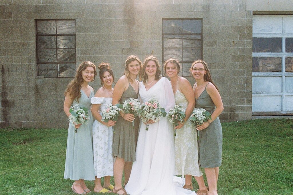 Film photo of a bride with her 5 bridesmaids standing together and smiling at the camera in front of a brick wall, taken by a documentary wedding photographer