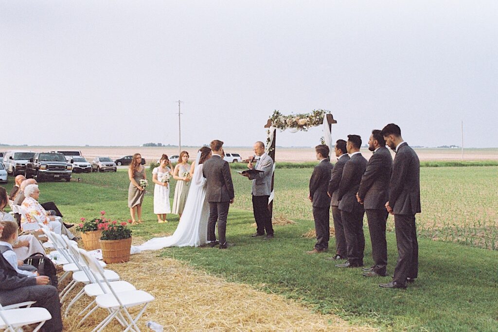 Film photo of a bride and groom standing holding hands in front of their officiant during their wedding ceremony with guests seated and their wedding parties on either side of them