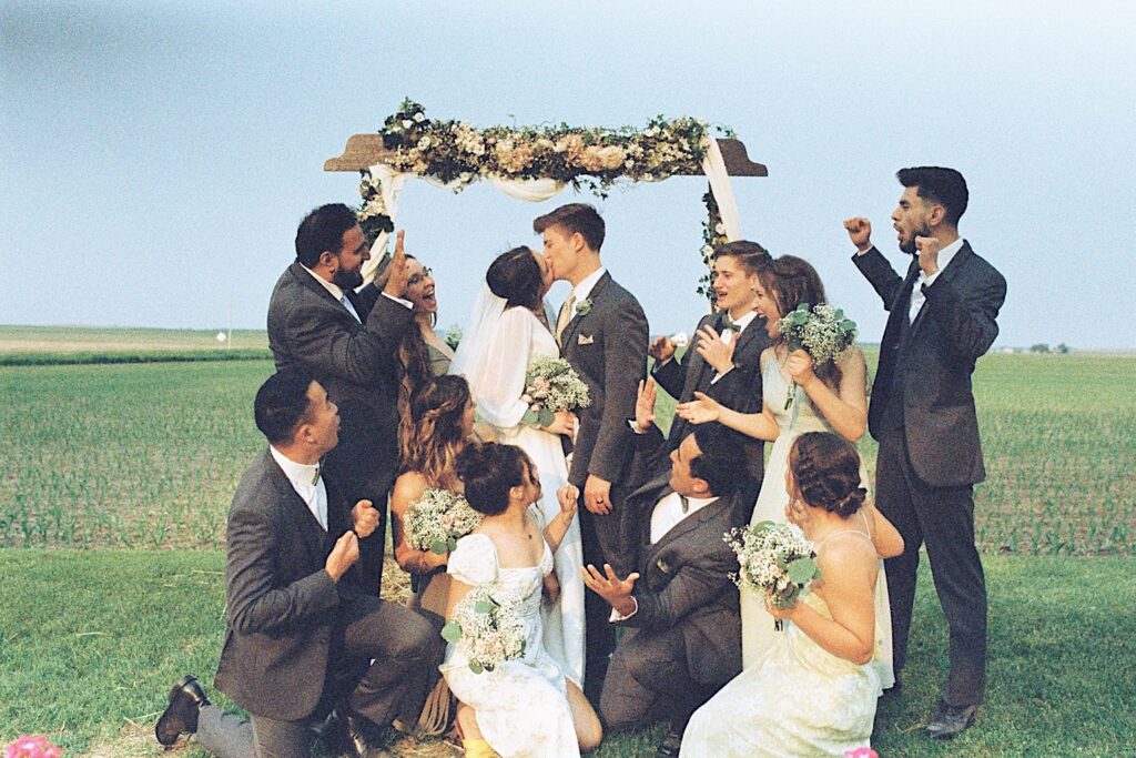 Film photo of a bride and groom kissing under a wooden arch in a field with their wedding parties surrounding them and cheering them on, taken by a documentary wedding photographer