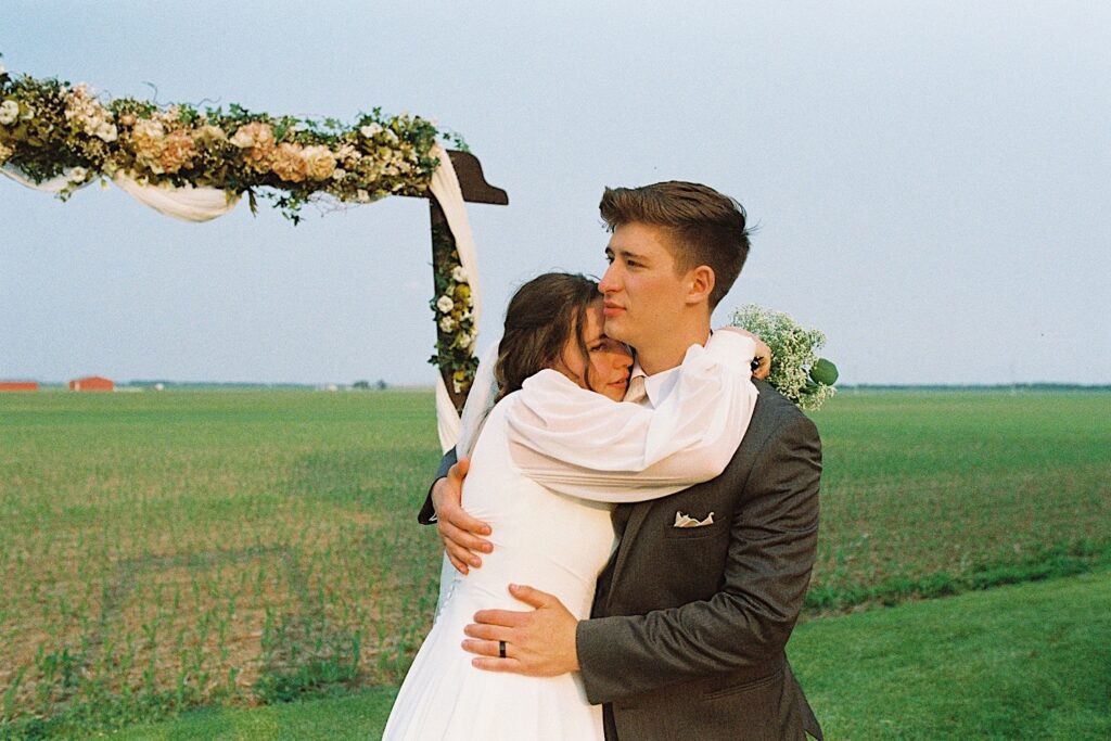 Film photo of a bride and groom embracing underneath a flower covered wooden arch in a cornfield, photographed by a documentary wedding photographer