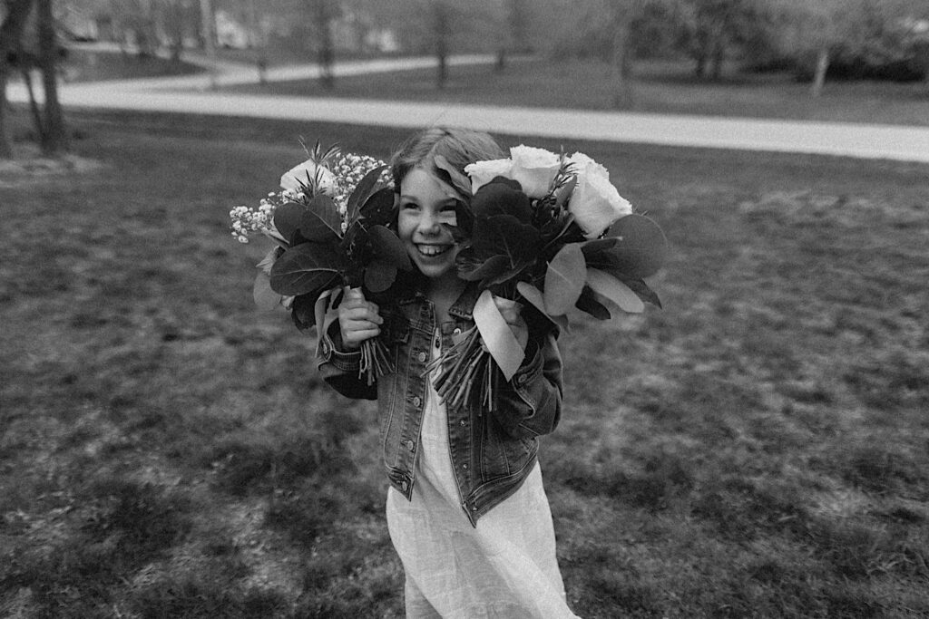 Black and white photo of a young girl smiling in a yard as she holds up 2 bouquets and sticks her face between the two