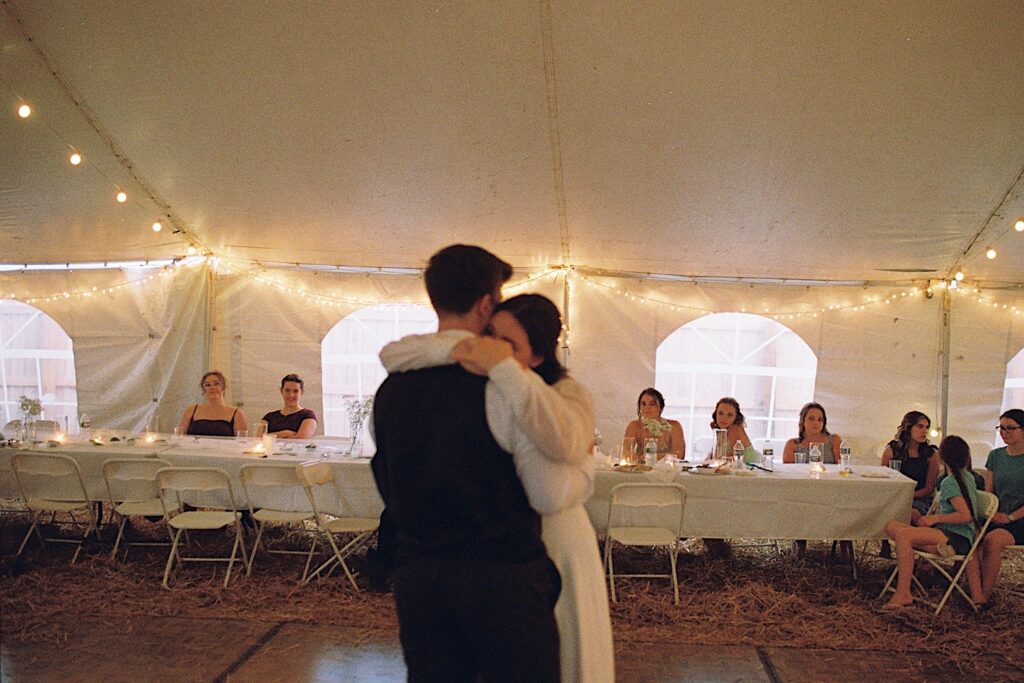 Film photo of a bride and groom as they embrace during their first dance under a tent as guests of their wedding sit and watch, taken by a documentary wedding photographer