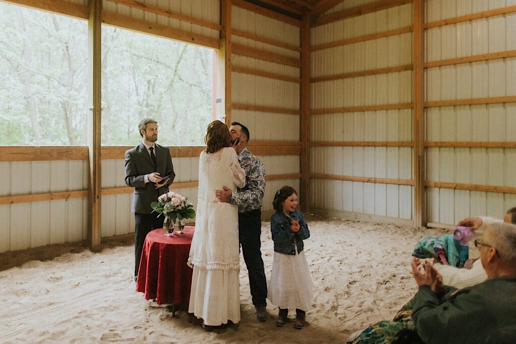 A bride and groom kiss as their daughter smiles beside them during their backyard wedding ceremony in their barn in Springfield Illinois